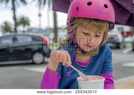 Young Girl In Pink Outfit And Pink Helmet Riding A Kick-scooter Sitting At The Outside Cafe Table Wi