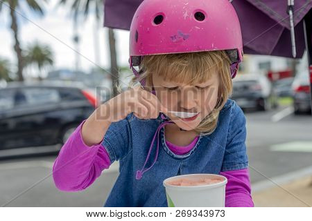 Young Girl In Pink Outfit And Pink Helmet Riding A Kick-scooter Sitting At The Outside Cafe Table Wi