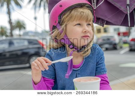 Young Girl In Pink Outfit And Pink Helmet Riding A Kick-scooter Sitting At The Outside Cafe Table Wi