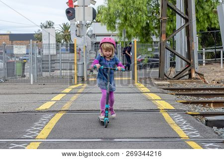 Young girl in pink outfit and pink helmet riding a kick-scooter across the railway line