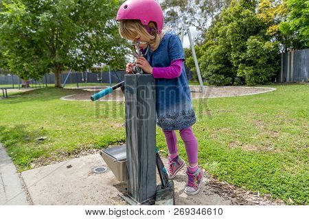 Young girl in pink outfit and pink helmet riding a kick-scooter in the park having a drink from foun