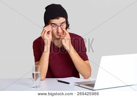 Photo Of Tired Man Rubbs Eyes, Wears Glasses, Has Pain In Eyes After Long Work At Laptop Computer, W