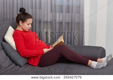Sideways Shot Of Pregnant Young Woman Reads Book, Rests At Home, Sits At Couch, Dressed In Red Sweat