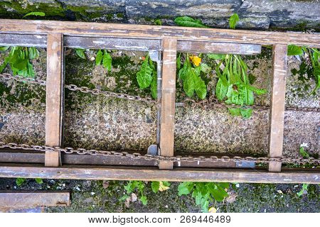 An Old Wooden Ladder Stepladder With Rusty Large Chains Lies On A Cement Surface Interspersed With S