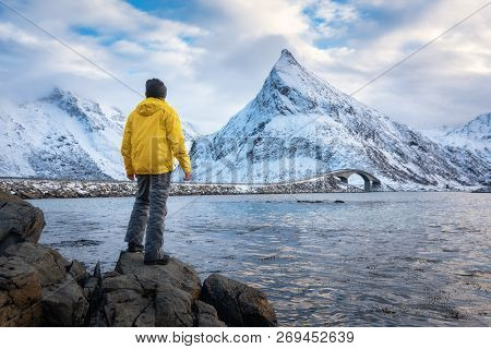 Sporty Man In Yellow Jacket Standing On The Stone On Seacoast Against Snowy Mountains And Cloudy Sky