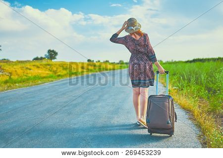 Pretty Female Hitchhiker In Summer Dress And Hat Hitchhiking With Suitcase. Cute Woman Walking On Th