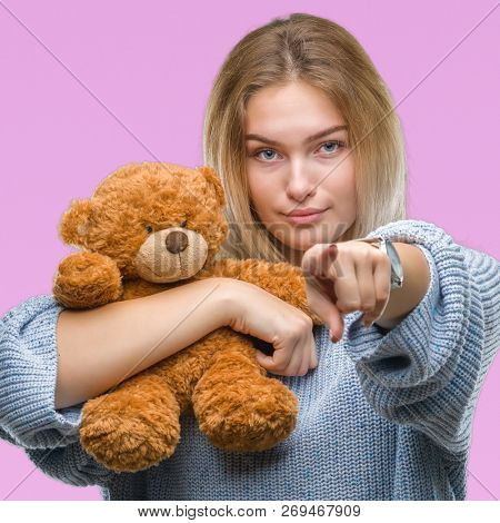 Young caucasian woman holding cute teddy bear over isolated background pointing with finger to the camera and to you, hand sign, positive and confident gesture from the front
