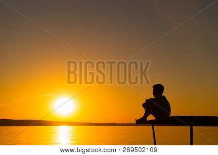 Silhouette Of A Little Boy Sitting On The Bridge In River At The Sky Sunset. A Lonely Boy Sits Quiet