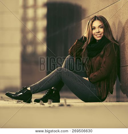 Happy young woman sitting at the wall in city street  Stylish fashion model wearing brown leather fringe suede jacket and dark blue jeans