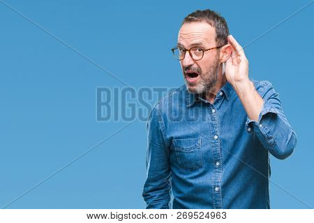 Middle age hoary senior man wearing glasses over isolated background smiling with hand over ear listening an hearing to rumor or gossip. Deafness concept.