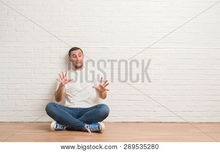 Young caucasian man sitting on the floor over white brick wall afraid and terrified with fear expression stop gesture with hands, shouting in shock. Panic concept.