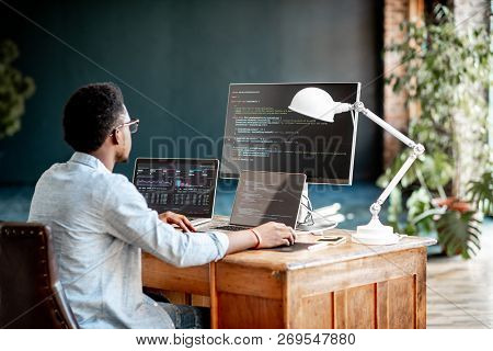 Young African Male Programmer Writing Program Code Sitting At The Workplace With Three Monitors In T