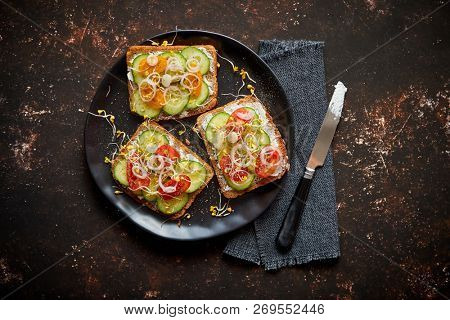 Vegetable toasts. Healthy toasts with cucomber, cherry tomatoes, crumbled feta cheese and radish sprouts on a black plate. Table top view. Dark rusty background.