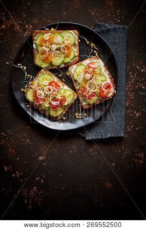 Vegetable toasts. Healthy toasts with cucomber, cherry tomatoes, crumbled feta cheese and radish sprouts on a black plate. Table top view. Dark rusty background.