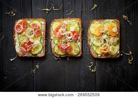 Row of three healthy toasts with onion, cucomber, cherry tomatoes, crumbled feta cheese and radish sprouts on a black plate. Table top view on black wooden background