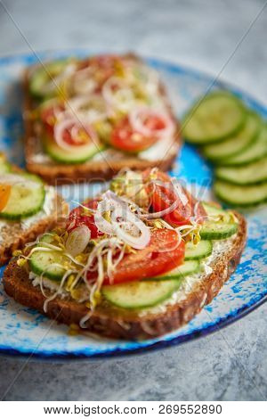 Plate with healthy toasts with cucomber, cherry tomatoes, crumbled feta cheese and radish sprouts on a black plate. Table top view. Gray stone background.