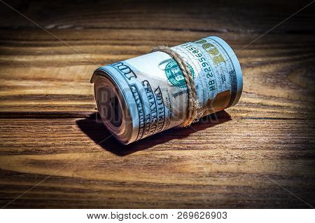 A Roll Of Dollars On A Wooden Textured Table In The Dark Illuminated By A Ray Of Light.
