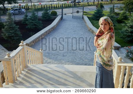 Woman in tatar head shawl stands on stairs outdoor.