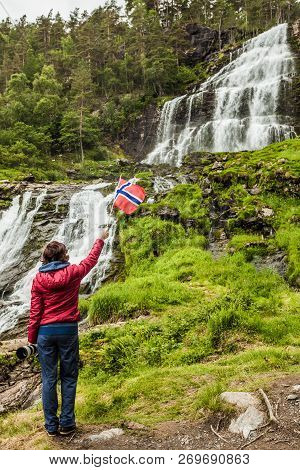 Tourist Woman With Norwegian Flag At Svandalsfossen In Norway, Powerful Waterfall In Norwegian Mount