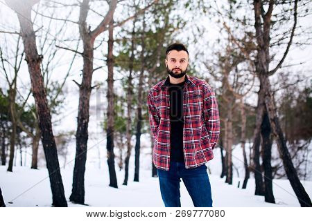 Portrait Of Handsome Bearded Young Man In Red Plaid Shirt Standing In Winter Snowy Forest. Stylish M