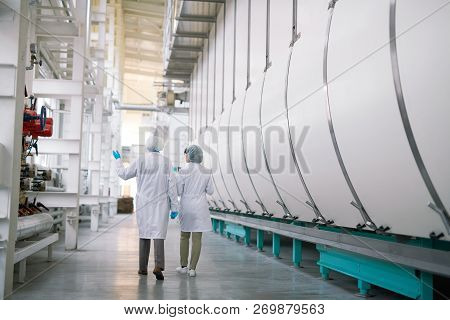 Back View Full Length Portrait Of Two Factory Workers Wearing White Coats Crossing Production Worksh