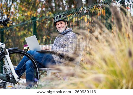 A Senior Man With Electrobike Sitting On A Bench Outdoors In Town, Using Laptop.