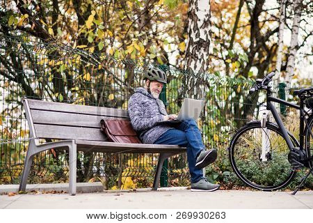 A Senior Man With Electrobike Sitting On A Bench Outdoors In Town, Using Laptop.