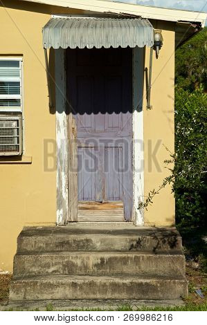 View Of Entrance With Rotted Door And Stained Cement Stairs And Weathered Awning On Abandoned Roadsi