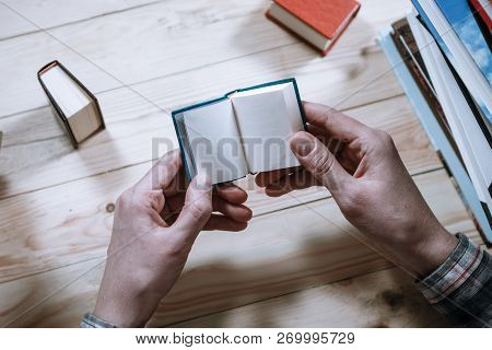 A Man Reading A Miniature Book Among The Big Books In The Library At The Wooden Table.