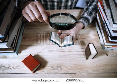 A Man Reading A Miniature Book Among The Big Books In The Library At The Wooden Table.