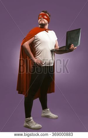 Super Hero Man Holds A Laptop Looking Up Thoughtfully. Business Man In Red Superhero Costume Using C