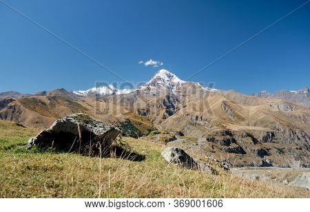 Snow Covered Mountain Kazbeg With Gergeti Trinity Church In The Foreground With Blue Sky Above, Step
