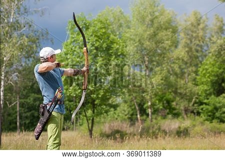 A Man In A Blue T-shirt And Cap Shoots An Archery. He Trains In The Woods.