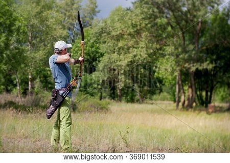 A Man In A Blue T-shirt And Cap Shoots An Archery. He Trains In The Woods.