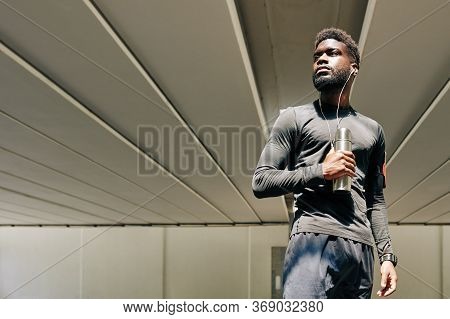 Portrait Of Confident Serious Sportsman With Small Thermos Listening To Music In Earbuds And Looking