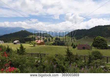 Country View, With Cabins And Barns At The Foot Of The Mountains. Cloudy Blue Sky. Lawns And Forests