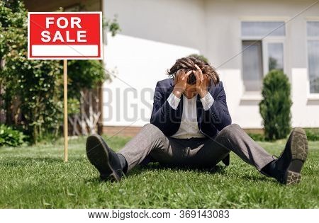 Depressed Real Estate Agent Sitting On Grass In Front Of House For Sale, Outside