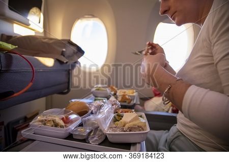 Women Passenger Of Aircraft Seats In Seat And Eats Tasty Hot Inflight Meal On A Folding Table. In Th