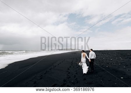 A Wedding Couple Is Walking Along The Black Beach Of Vic. Sandy Beach With Black Sand On The Shores 
