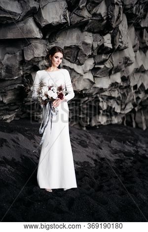 Destination Iceland Wedding. The Bride In A White Silk Dress With A Bouquet In Her Hands, Stands On 
