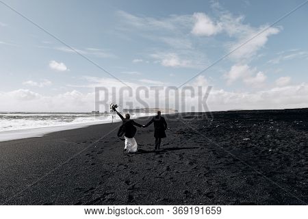Destination Iceland Wedding. The Wedding Couple Runs Along The Sandy Black Beach Of Vik, Near The Ba