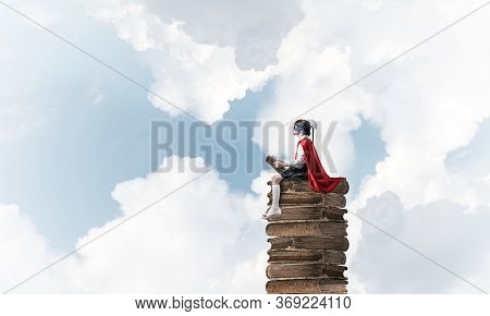 Little Child In Mask And Cape Sittting On Pile Of Old Books In Sky And Reading