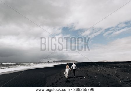 A Wedding Couple Is Walking Along The Black Beach Of Vic. Sandy Beach With Black Sand On The Shores 
