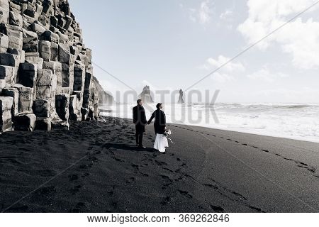 Destination Iceland Wedding. A Wedding Couple Walks Along The Sandy Black Beach Of Vik, Near The Bas