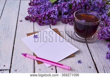 Cup Of Tea With Lilac Flowers On A Wooden White Background. Nearby Lies A Sheet Of Paper, And A Pink