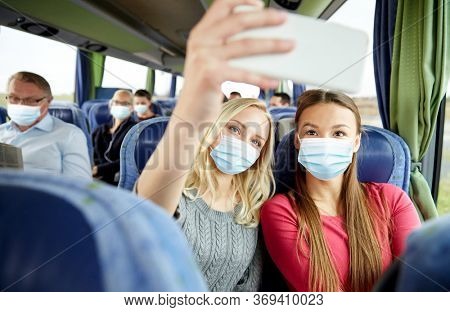 tourism, pandemic and health care concept - young women or friends wearing face protective medical mask for protection from virus disease in travel bus taking selfie by smartphone