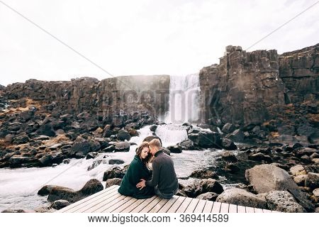 A Guy And A Girl Are Sitting Embracing On A Bridge Near Ehsaraurfoss Waterfall, Ehsarau River, Natio