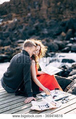 A Guy Hugs A Girl Artist Who Paints A Picture In The Album With Watercolors. Near Ehsaraurfoss Water