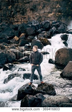 A Guy In A Black Knitted Sweater Stands On Rocks Near Ehsaraurfoss Falls, Ehsarau River, National Pa