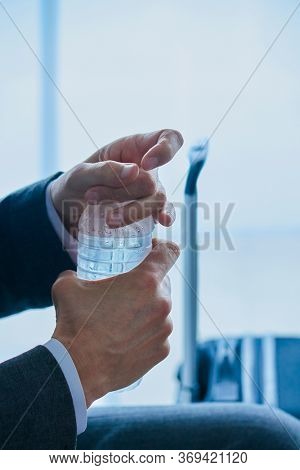 closeup of a young businessman, in a gray suit, sitting in the waiting hall of an airport or a train station, opening a bottle of cold water next to his suitcase